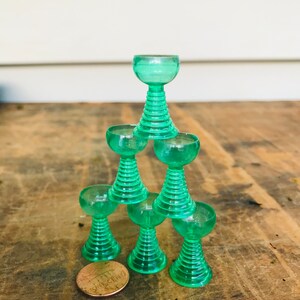 May include: A stack of six green glass goblets with a fluted design. The goblets are stacked in a pyramid shape, with the smallest goblet on top. A penny is in the foreground.