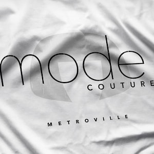 May include: A black and white image of a white fabric with the words "mode COUTURE METROVILLE" printed on it in a minimalist font. The word "mode" is in a large, stylized font, with "COUTURE" and "METROVILLE" in a smaller font.