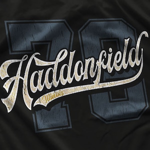 Könnte beinhalten: Ein schwarzes T-Shirt mit einem Vintage-Design, das den Text "Haddonfield" in einer weißen Schriftart mit goldenen Akzenten zeigt. Die Nummer "70" befindet sich im Hintergrund in einem blau-weißen Farbschema.