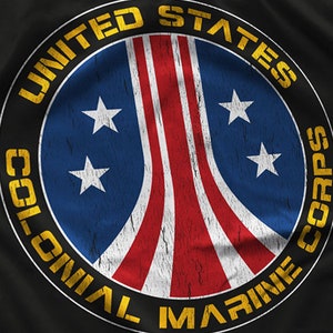Op de afbeelding: Cirkelvormig embleem met de tekst "UNITED STATES COLONIAL MARINE CORPS" in geel. Het embleem heeft een blauwe achtergrond met vier witte sterren en rood-witte strepen, met een verweerde, vintage look.