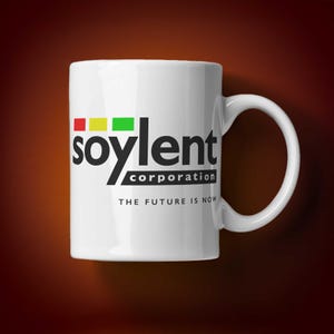 Op de afbeelding: Witte keramische mok met de tekst "soylent corporation" in zwart, met rode, gele en groene vierkanten boven het woord "soylent". De tekst "THE FUTURE IS NOW" staat eronder.