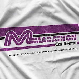 Könnte beinhalten: Ein weißes T-Shirt mit einem lila und schwarzen Logo, auf dem "Marathon Car Rental" steht. Das Logo zeigt ein stilisiertes "M" und den Text "Wählen Sie zwischen Modellen von Datsun, Toyota, Mustang und Buick".