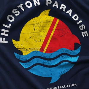 May include: A navy blue t-shirt with a white circle graphic. The graphic features a yellow sun with a red and yellow stripe, a blue wave, and a white shark fin. The text "FHLOST PARADISE" is written in a white circle around the graphic.