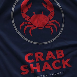 Pode incluir: Uma t-shirt azul-marinho com um gráfico de caranguejo vermelho dentro de uma borda circular com textura desgastada. Abaixo do círculo, o texto CRAB SHACK está impresso em vermelho, seguido pelo texto branco menor CAMDEN COUNTY em fonte sem serifa.