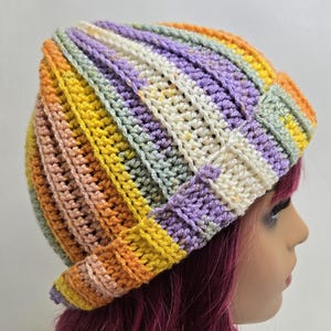 May include: A multi-coloured, striped crochet beanie in shades of yellow, orange, pink, green, purple, and white. The hat has a ribbed stitch and a folded brim. This handmade hat is a cosy accessory.