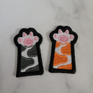 Könnte beinhalten: Zwei gestickte Katzenpfoten-Aufnäher. Ein Aufnäher zeigt einen grau-weißen Pfotenabdruck mit einem rosa Ballen, der andere einen orange-weißen Pfotenabdruck mit einem rosa Ballen. Beide Aufnäher haben einen schwarzen Rand.