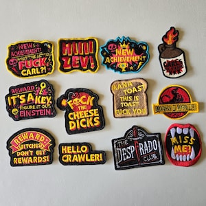 May include: A collection of eleven embroidered patches featuring various designs and text, including phrases like New Achievement, It's a Key, Fuck the Cheese Dicks, Mana Toast, Mongo U Appalled, Ditches Don't Get Rewards, Hello Crawler, and The Desperado Club.
