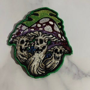 Op de afbeelding: Een geborduurde patch met drie paddenstoelen met schedels aan de basis. De paddenstoelen zijn paars, rood en groen met witte stippen. De schedels zijn wit met zwarte ogen en tanden. De patch is omrand met groen.