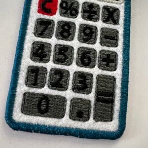 May include: A blue and white embroidered patch of a calculator with black numbers and symbols. The calculator has a red button in the top left corner.