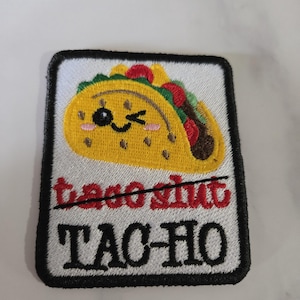 Puede incluir: Parche bordado con un diseño de taco de dibujos animados. El taco es amarillo con rellenos rojos, verdes y marrones. El texto "TAC-HO" está debajo del taco, con "taco slut" tachado.