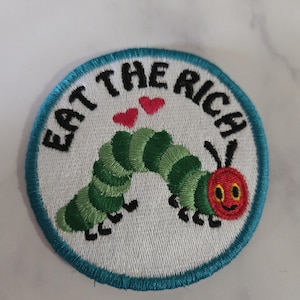 Może przedstawiać: Haftowana naszywka z białym tłem i turkusową obwódką. Naszywka przedstawia zieloną gąsienicę z czerwoną głową i czarnym napisem "EAT THE RICH". Dwa czerwone serca znajdują się nad gąsienicą.