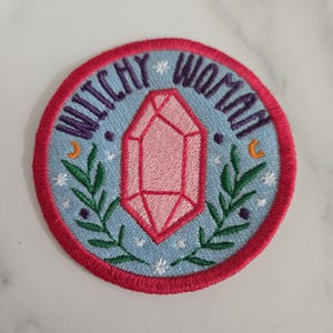 May include: A round, embroidered patch with a pink crystal, green leaves, and the text "Witchy Woman" in purple and white lettering on a blue background.