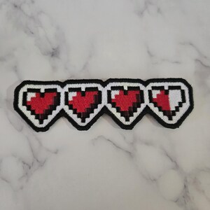 May include: A pixelated patch featuring four red hearts with black outlines. The hearts are in a row, with the last heart partially white. The patch has a black border and is likely for a video game enthusiast.