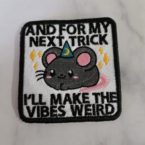 Könnte beinhalten: Gestickter Aufnäher mit einer schwarzen Maus, die einen Zaubererhut, rosa Ohren und einen rosa Schwanz trägt. Der Text lautet "AND FOR MY NEXT TRICK I'LL MAKE THE VIBES WEIRD" mit gelben Sternen-Akzenten. Der Aufnäher ist quadratisch mit einem schwarzen Rand.