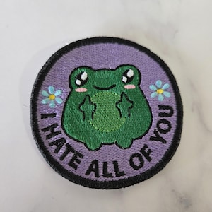 Op de afbeelding: Een ronde geborduurde patch met een paarse achtergrond met een groene kikker met roze wangen en grote ogen. De kikker steekt zijn middelvingers op. Twee kleine blauwe bloemen staan aan de zijkanten. De tekst I HATE ALL OF YOU is geborduurd.