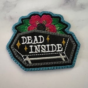 May include: Embroidered patch with a black coffin with the text "DEAD INSIDE" in white. The coffin is decorated with a pink flower and green leaves. There are gold stars on the coffin.