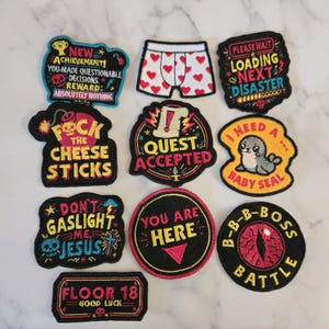 May include: A collection of ten embroidered patches featuring various designs including a pair of heart-patterned boxers, a seal, a red eye, and text such as New Achievement, Loading Next Disaster, Quest Accepted, and Floor 18.