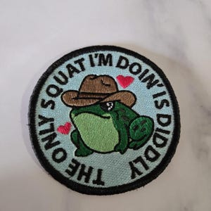 May include: Round embroidered patch featuring a green frog wearing a brown cowboy hat. The patch has the text "ONLY SQUAT I'M DOIN' IS DIDDLY" in a circular pattern around the frog, with two pink hearts.