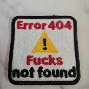May include: Embroidered patch with a white background, black border, and the text "Error 404 Fucks not found." A yellow triangle with an exclamation point is centered above the text. The text is in red and black.