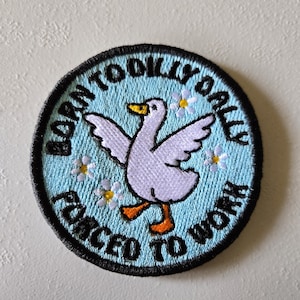 May include: A circular embroidered patch with a light blue background featuring a white goose with orange feet and beak. The text BORN TO DILLY DALLY FORCED TO WORK is stitched in black around the border, accented by small white flowers.