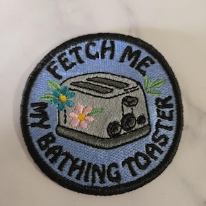 Puede incluir: Parche bordado con fondo azul claro y borde negro. El parche presenta una tostadora gris con el texto "FETCH ME MY BATHING TOASTER" en negro. Flores y hojas coloridas están bordadas alrededor de la tostadora.