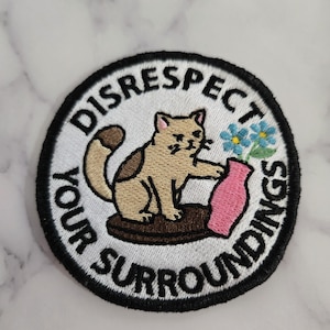 Könnte beinhalten: Gestickter Aufnäher mit einer Katze, die nach einer rosa Vase mit blauen Blumen greift. Der Aufnäher trägt die Worte "DISRESPECT YOUR SURROUNDINGS" in einem kreisförmigen Design mit schwarzem Rand.