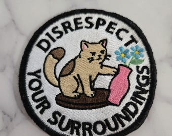 May include: Embroidered patch with a cat reaching for a pink vase with blue flowers. The patch has the words "DISRESPECT YOUR SURROUNDINGS" in a circular design, with a black border.