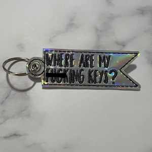May include: A silver iridescent keychain with a black embroidered phrase "Where are my [censored] keys?"