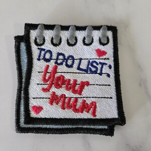 May include: A square embroidered patch shaped like a notepad with a black border and grey spiral binding at the top. The white fabric features black horizontal lines and the text TO DO LIST: Your mum in blue and red thread, with two small red hearts.