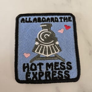 May include: Embroidered patch with a light blue background, featuring a black and gray train illustration. The text "ALL ABOARD THE HOT MESS EXPRESS" is displayed in black. Pink and red heart accents are also present.