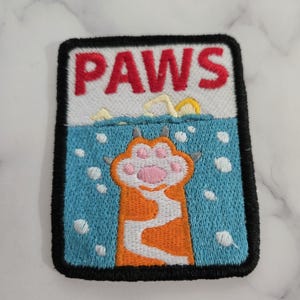 Könnte beinhalten: Gestickter Aufnäher mit dem Wort "PAWS" in roten Buchstaben über einem Cartoon-Katzenpfoten-Design. Die Pfote ist orange und weiß, mit einem rosa Pfotenballen. Der Hintergrund ist blau mit weißen Blasen und einem gelb-weißen Wellenmuster.