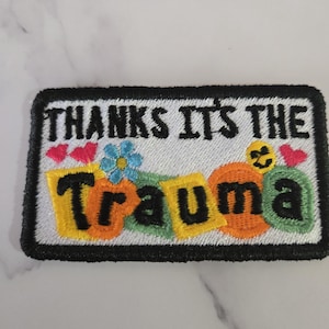 May include: Embroidered patch with the text "THANKS IT'S THE Trauma" in black letters. The patch features colorful accents, including hearts, flowers, and orange and yellow shapes. The patch has a black border.