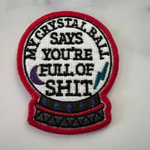 May include: A red embroidered patch shaped like a crystal ball with the text "My Crystal Ball Says You're Full Of Shit" in black. The crystal ball has a crescent moon and lightning bolt inside.