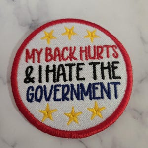 Puede incluir: Parche bordado con el texto "MY BACK HURTS & I HATE THE GOVERNMENT". El parche tiene un fondo blanco, un borde rojo y detalles de estrellas amarillas. El texto es en rojo, negro y azul.