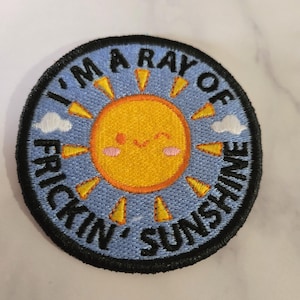 May include: Round embroidered patch with a smiling sun design. The sun is yellow and orange, surrounded by yellow rays and a light blue background. The text "I'M A RAY OF FRICKIN' SUNSHINE" is embroidered in black.
