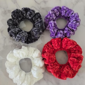 May include: Four plush scrunchies in a variety of colors: dark gray, purple, red, and white. The scrunchies are made of a soft, velvety material and are arranged on a light-colored surface. These hair accessories are perfect for adding a touch of color and texture to any hairstyle.