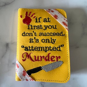 Puede incluir: Funda de pasaporte amarilla con el texto bordado "if at first you don't succeed, it's only 'attempted' Murder". Presenta una huella de mano roja, un cuchillo y una cinta con salpicaduras rojas.