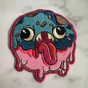 Pode incluir: Um remendo bordado com um monstro de desenho animado com uma língua grande para fora. O monstro tem olhos saltados e um corpo rosa e azul que pinga.