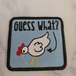 May include: Embroidered patch with a light blue background and a black border. The patch features a white chicken with a red comb and yellow legs. The text "GUESS WHAT?" is above the chicken.