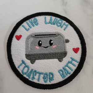 Könnte beinhalten: Gestickter Aufnäher mit weißem Hintergrund, schwarzem Rand und den Worten "Live Laugh Toaster Bath" in Türkis. Ein grauer Toaster mit einem lächelnden Gesicht und rosa Wangen befindet sich in der Mitte, mit zwei roten Herzen. Ein lustiges, skurriles Design.