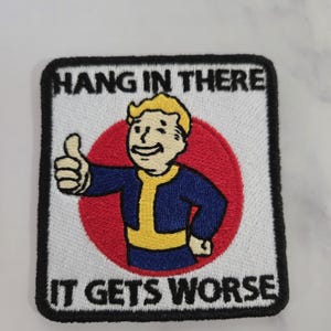 May include: A square embroidered patch with a white background and a black border. It features a cartoon character with yellow hair wearing a blue and yellow jumpsuit, giving a thumbs up in front of a red circle. The text reads HANG IN THERE IT GETS WORSE.