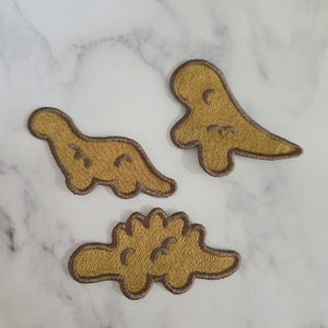 May include: Three embroidered iron-on patches featuring cartoon dinosaurs in a light brown color. The dinosaurs are a stegosaurus, a brachiosaurus, and a triceratops.
