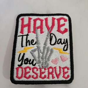 May include: Embroidered patch with the text "HAVE The Day You DESERVE" in red and black lettering. The design includes a gray skeleton hand, yellow lightning bolts, and pink hearts on a white background.
