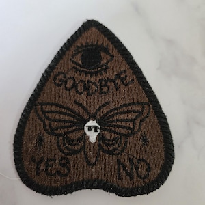 May include: Brown felt patch in the shape of a triangle with a black embroidered eye, butterfly, and the words "Goodbye", "Yes", and "No".
