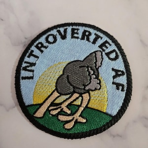 Op de afbeelding: Geborduurde ronde patch met de tekst "INTROVERTED AF". De patch toont een cartoon struisvogel met zijn hoofd naar beneden, tegen een blauwe lucht en gele zon, met een groen grasveld aan de onderkant.