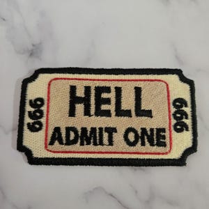 以下が含まれることがあります： 「HELL ADMIT ONE」の黒い文字が入ったチケット型の刺繍パッチ。 パッチは、赤いアクセントと両側に数字「666」が入ったベージュの背景です。 境界線は黒です。