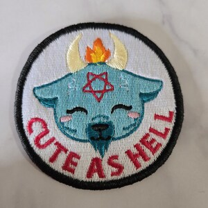 Może przedstawiać: Haftowana naszywka z jasnoniebieskim motywem kozy, czerwonym pentagramem, żółtymi rogami i płomieniem. Tekst "CUTE AS HELL" jest czerwony, z czarną obwódką i białym tłem.