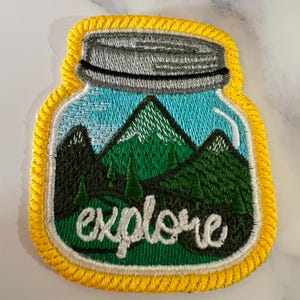 May include: A yellow embroidered patch with a mason jar design. The jar contains a green mountain scene with the word "explore" written in white on the bottom.