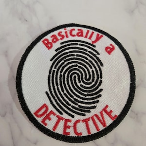 May include: Circular embroidered patch featuring a black fingerprint design on a white background. The words "Basically a DETECTIVE" are embroidered in red. The patch has a black border.