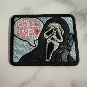 May include: Embroidered patch featuring a white ghost face mask with a black hooded robe, holding a blue phone. The ghost is saying "Call Me" in a pink speech bubble with a pink heart.